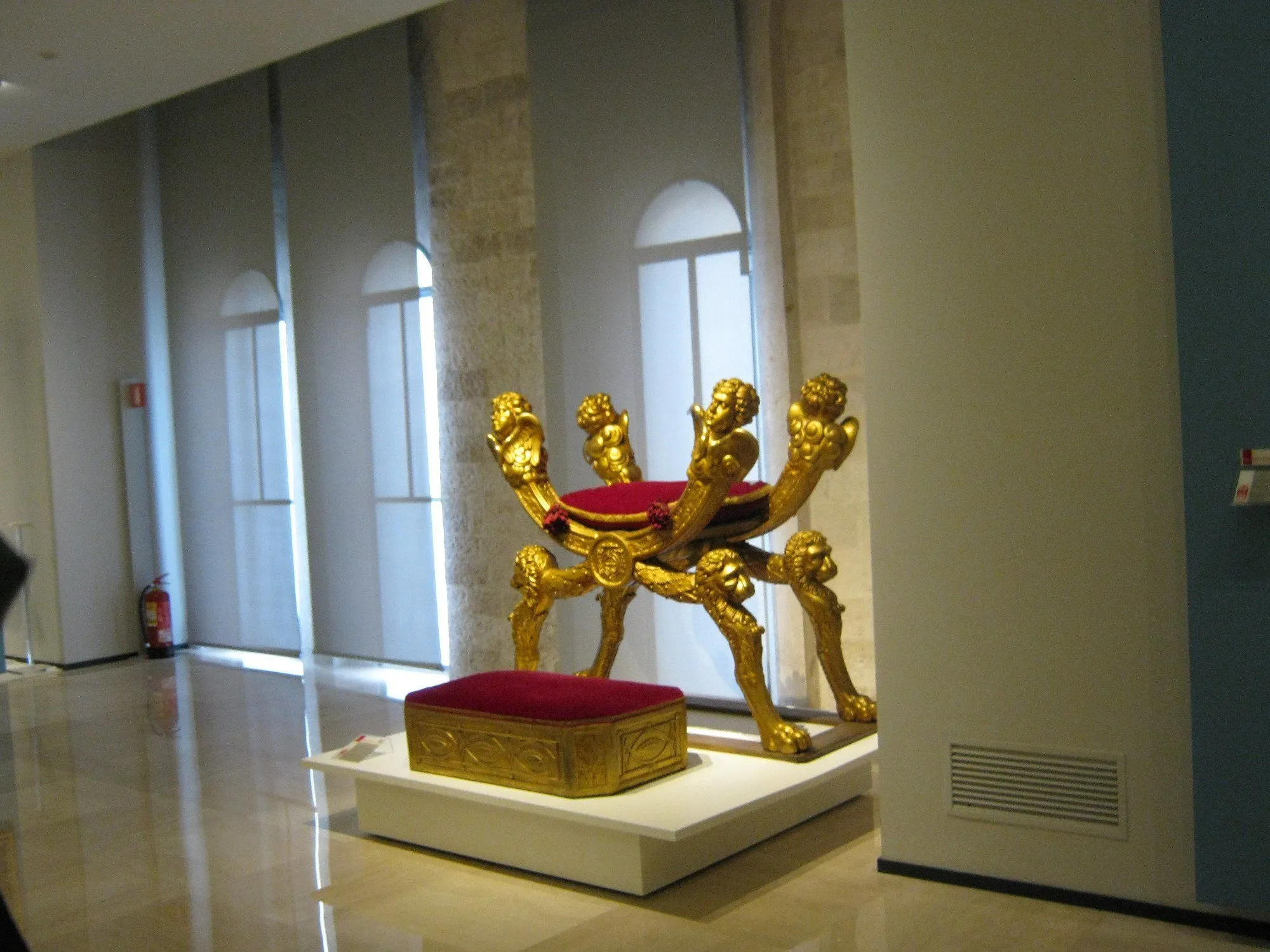 An editorial style image showcasing an ornate golden chair, possibly a throne, with a red velvet cushion and a matching footstool, displayed on a white pedestal in a well-lit indoor setting, likely a museum or gallery. The background features large windows with blinds and stone architectural elements.