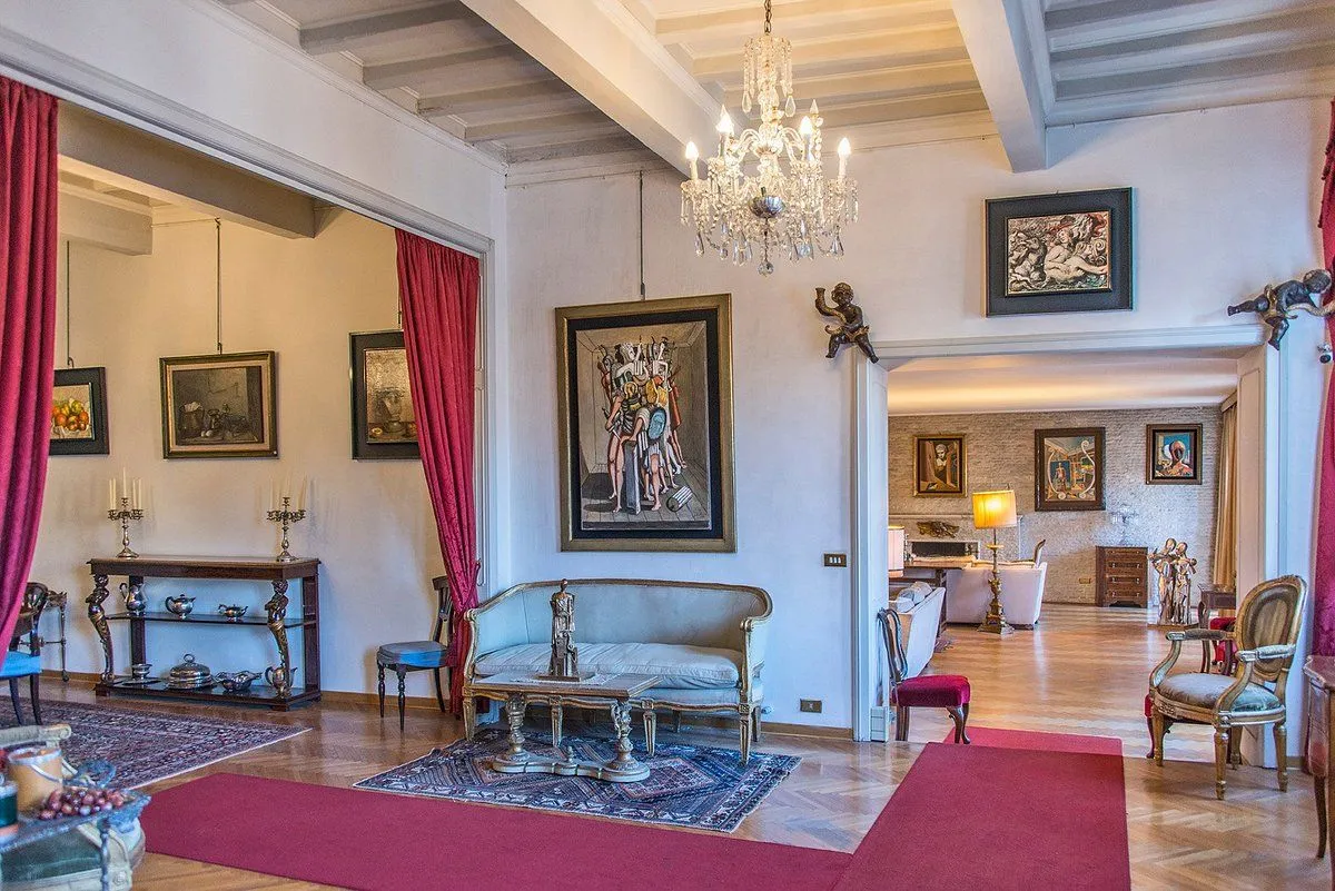 An elegant and ornate interior featuring classical furniture, a large chandelier, and a prominent painting. The space appears to be a formal living room or reception area, with a plush red carpet leading into the room and another room visible through an archway in the background.
