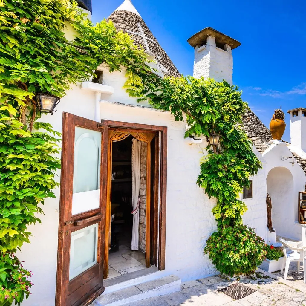 An editorial style image featuring a traditional Trullo house with white walls, a conical stone roof, and green climbing foliage under a clear blue sky.