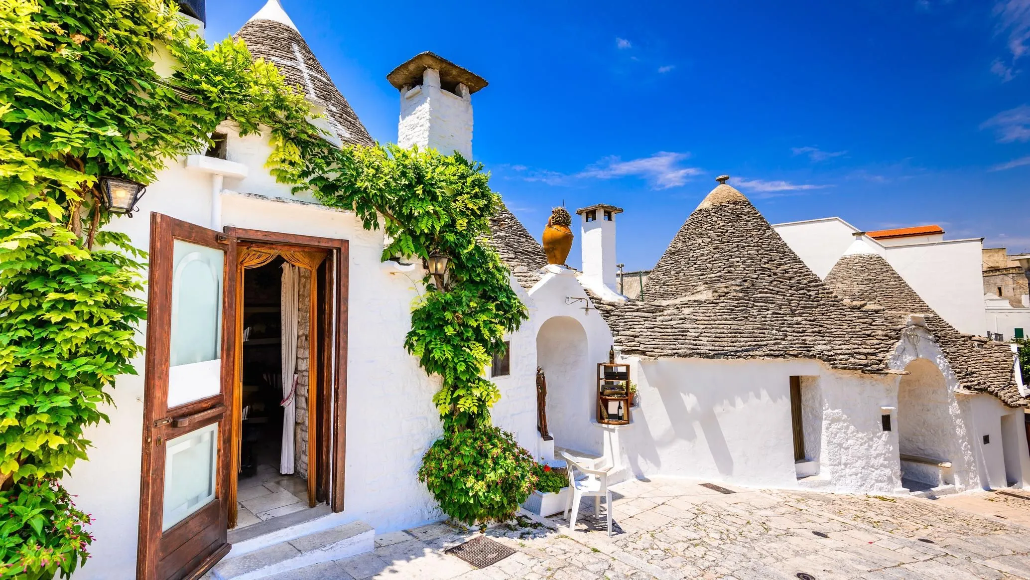 An editorial style image featuring a traditional Trullo house with white walls, a conical stone roof, and green climbing foliage under a clear blue sky.