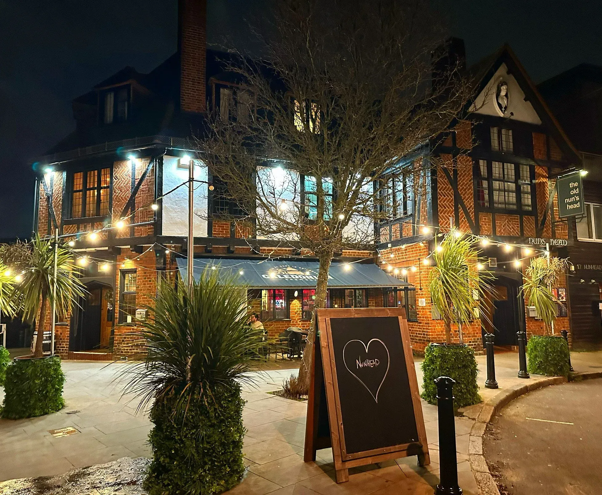 An inviting night-time view of a brick building, likely a restaurant or bar, with string lights illuminating its facade and an outdoor seating area. A tree stands in front, and a chalk A-frame sign is visible in the foreground.