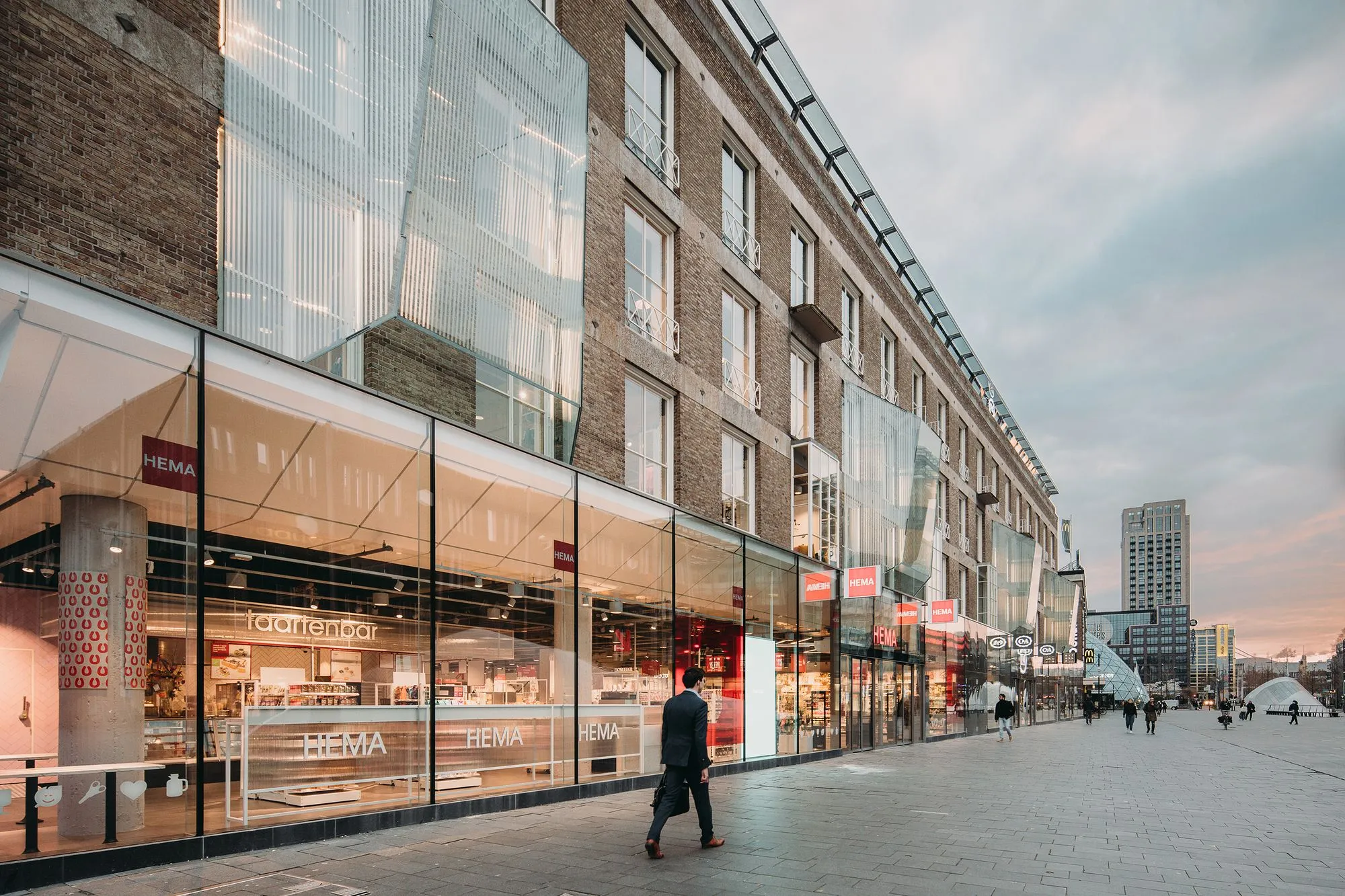 A well-composed and sharply focused photograph capturing a modern street scene. The image features a multi-story building with a blend of traditional brickwork and contemporary glass facades, under soft, even lighting. The colors are accurate, and key elements like the architecture and a walking pedestrian are in crisp focus, contributing to a high degree of photo-realism.