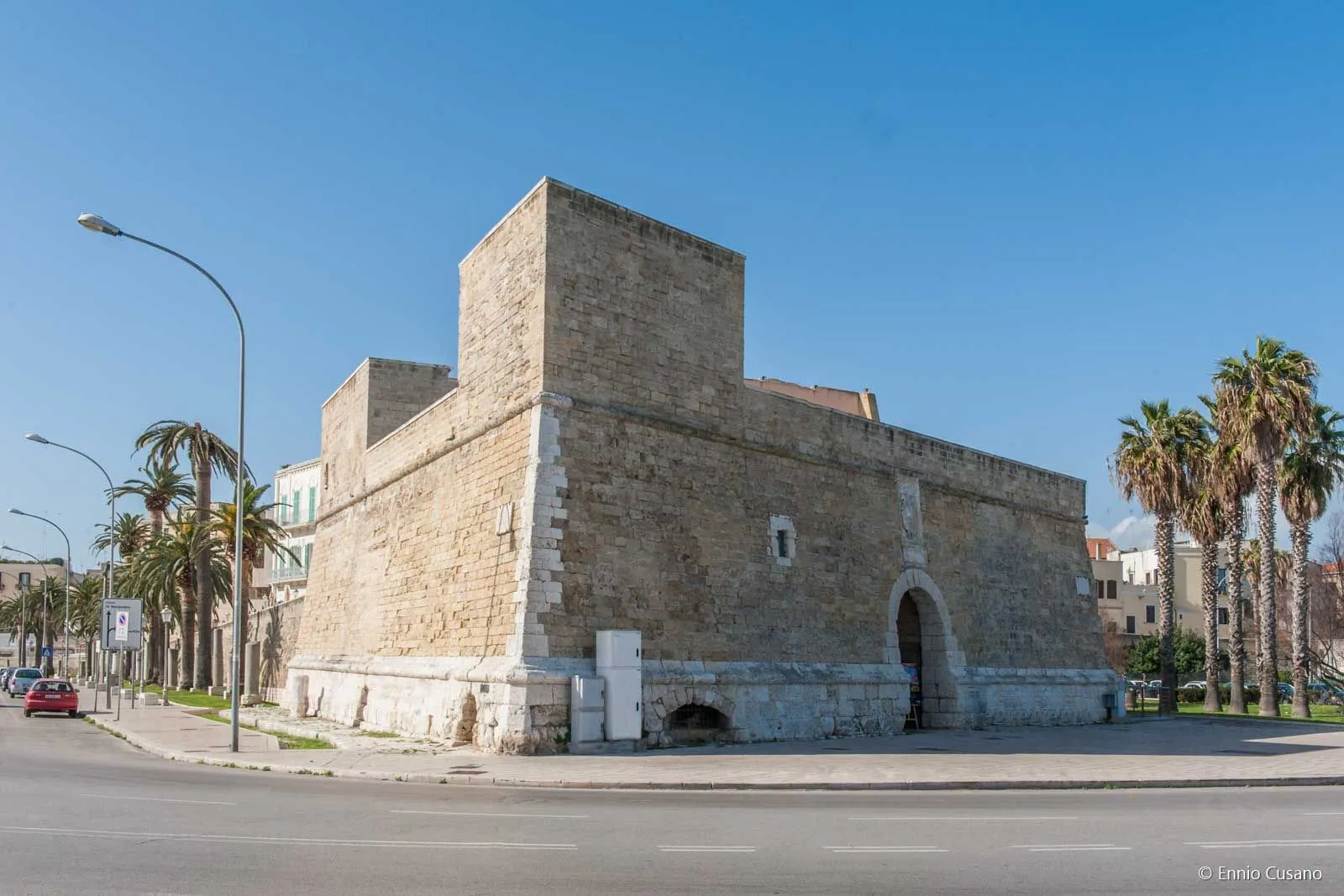 An exterior view of an ancient stone fortress or castle with a prominent square tower, situated by a road under a clear blue sky.