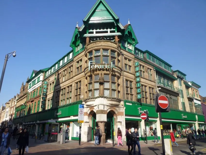 An exterior shot of the Fenwick department store building under clear skies. The building features a distinctive green roof and ornate stone facade. Several people and street signs are visible on the sidewalk in the foreground.