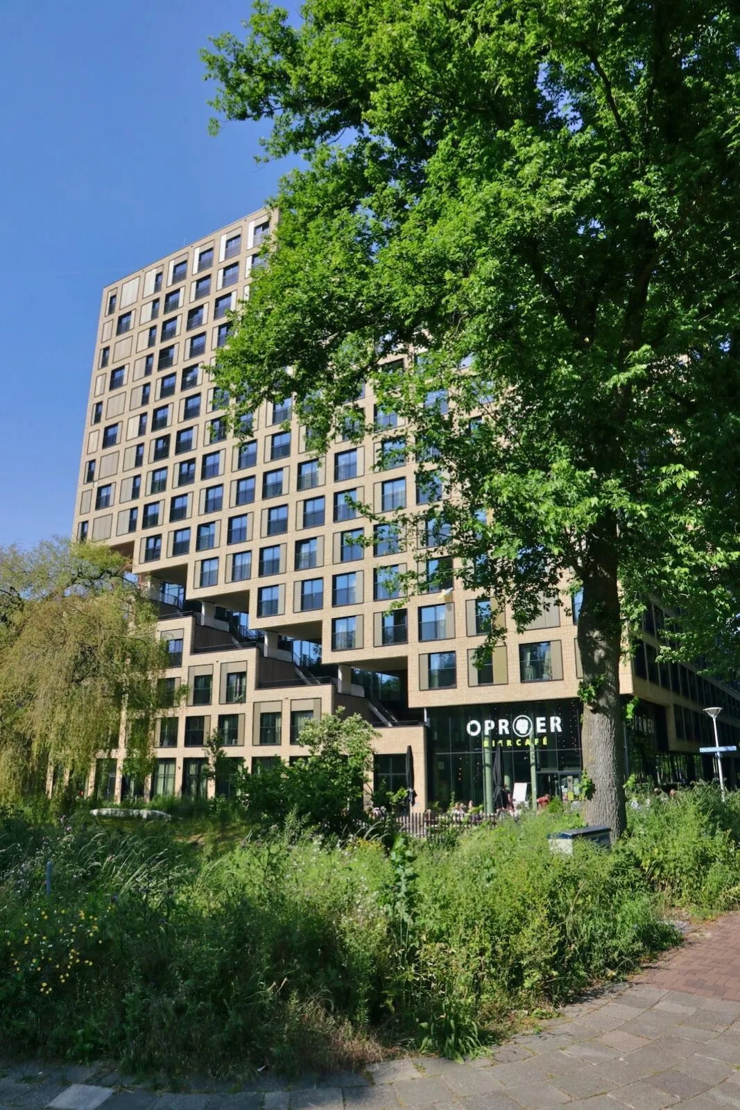 An exterior shot of a modern, angular building featuring a ground-floor establishment named "OPROER BIERCAFÉ", partially framed by lush green trees and foliage in the foreground under a clear blue sky.