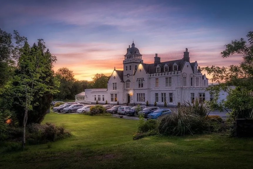 An impressive exterior shot of a large, white hotel building at sunset, featuring a vibrant sky, green lawns, and a parking area with cars. The image showcases the architectural details of the building under soft, warm light.