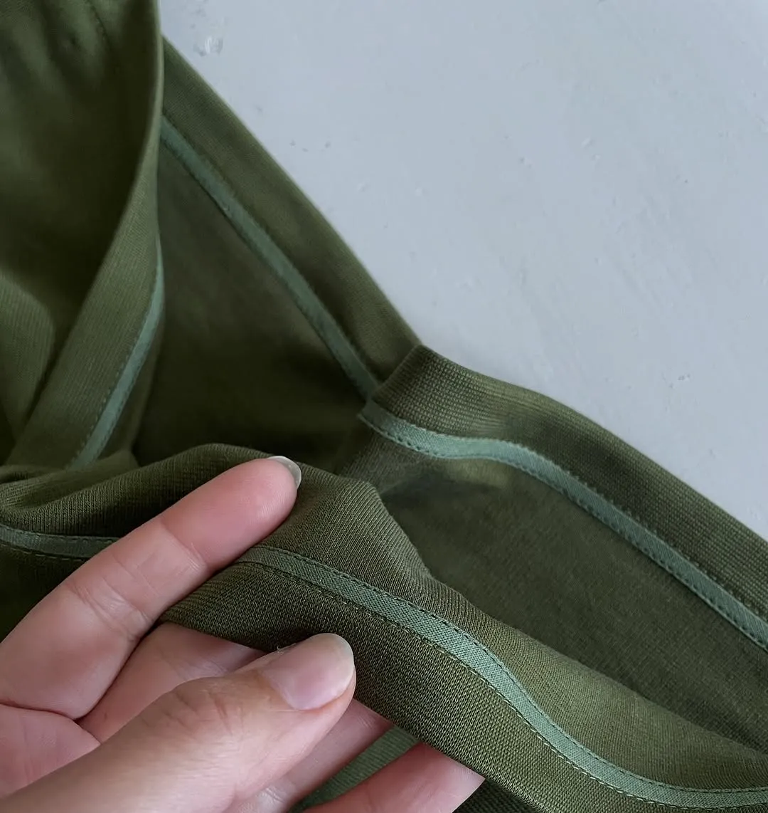A close-up image of a green fabric, possibly part of a garment, held by a hand, showcasing its texture, stitching, and a lighter green stripe detail against a plain white background.