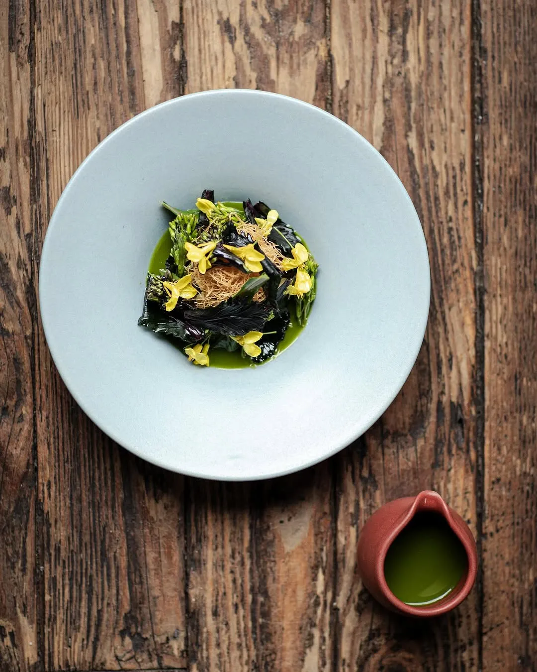This is a high-quality, professionally taken photograph of a gourmet dish. The composition is well-balanced, with the main dish thoughtfully placed on a muted blue plate against a rustic wooden table. The lighting is excellent, providing an even illumination that highlights the textures and colors of the food without harsh shadows. The image is very sharp, ensuring that all key elements, particularly the delicate greens, yellow flowers, and crispy threads, are in clear focus. The colors appear accurate and vibrant, contributing to the overall photo-realistic appeal of the dish. No unwanted elements are present, and the presentation is visually appealing.