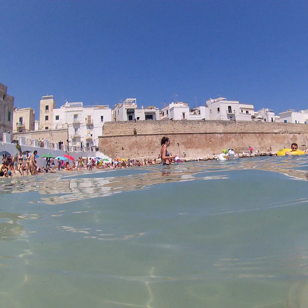 An editorial-style image capturing a sunny beach scene with a historic walled town in the background, featuring a unique split-shot perspective showing both above and below the water.