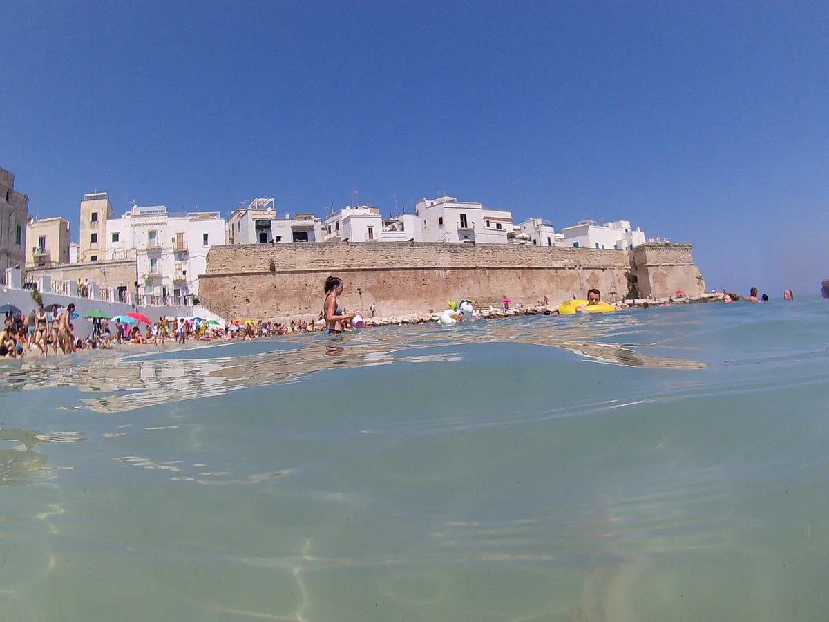 An editorial-style image capturing a sunny beach scene with a historic walled town in the background, featuring a unique split-shot perspective showing both above and below the water.