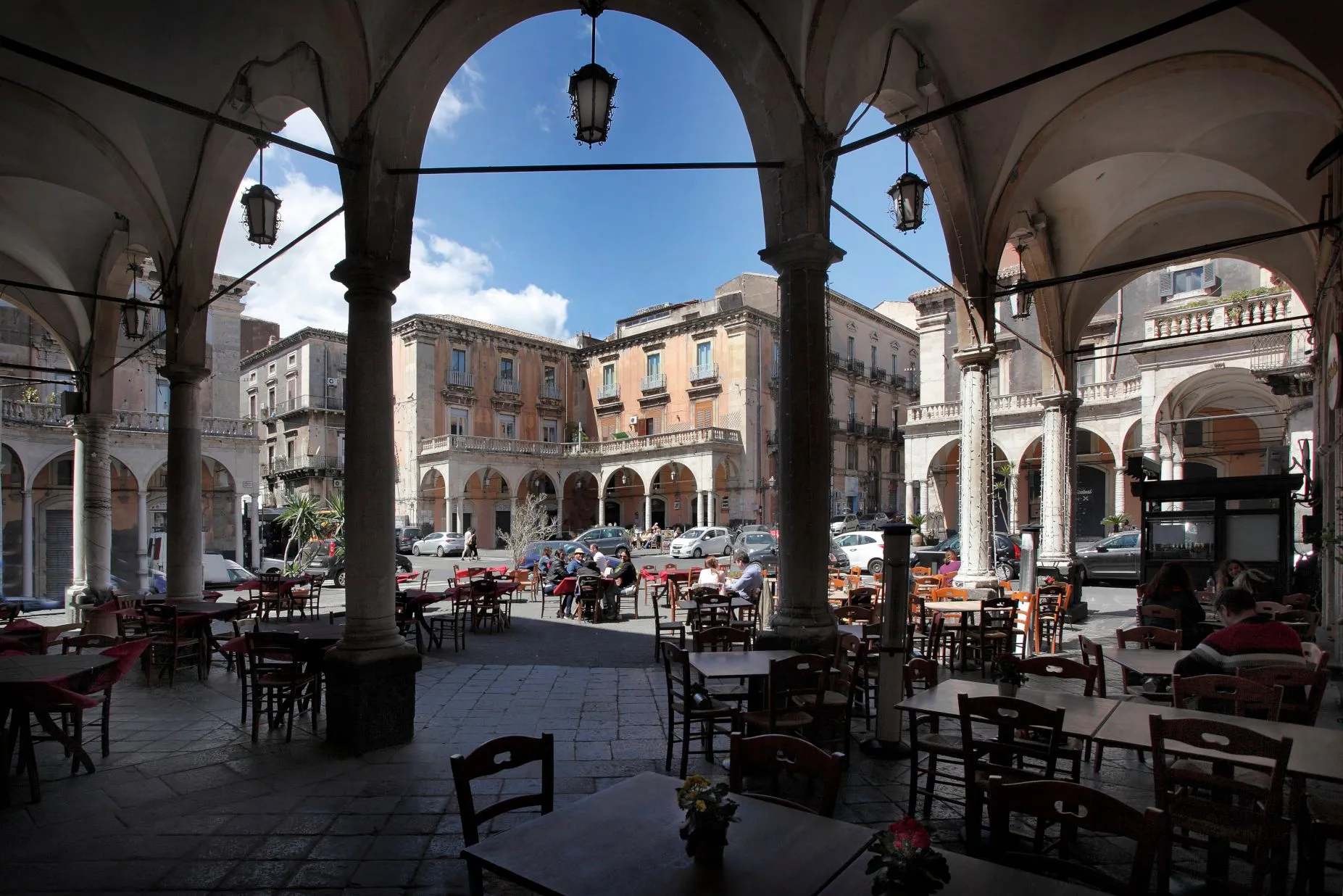 An editorial image showcasing a bustling city square framed by ancient stone arches, featuring outdoor cafe seating in the foreground and historic buildings with people and cars in the sunlit background.