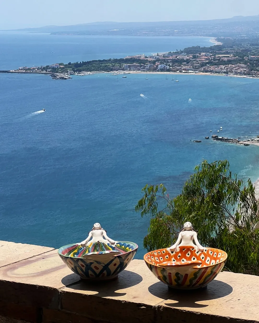 The image is well-composed, featuring unique decorative bowls in the foreground that frame a stunning seascape and distant coastline. It exhibits good overall sharpness and realistic, vibrant colors, particularly the deep blue of the sea.