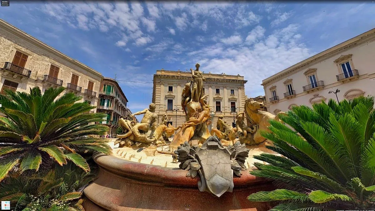 A low-angle, wide-angle shot of a grand stone fountain with multiple sculptures, set against a backdrop of historic European buildings under a clear blue sky with scattered clouds. Palm fronds are visible in the lower corners.