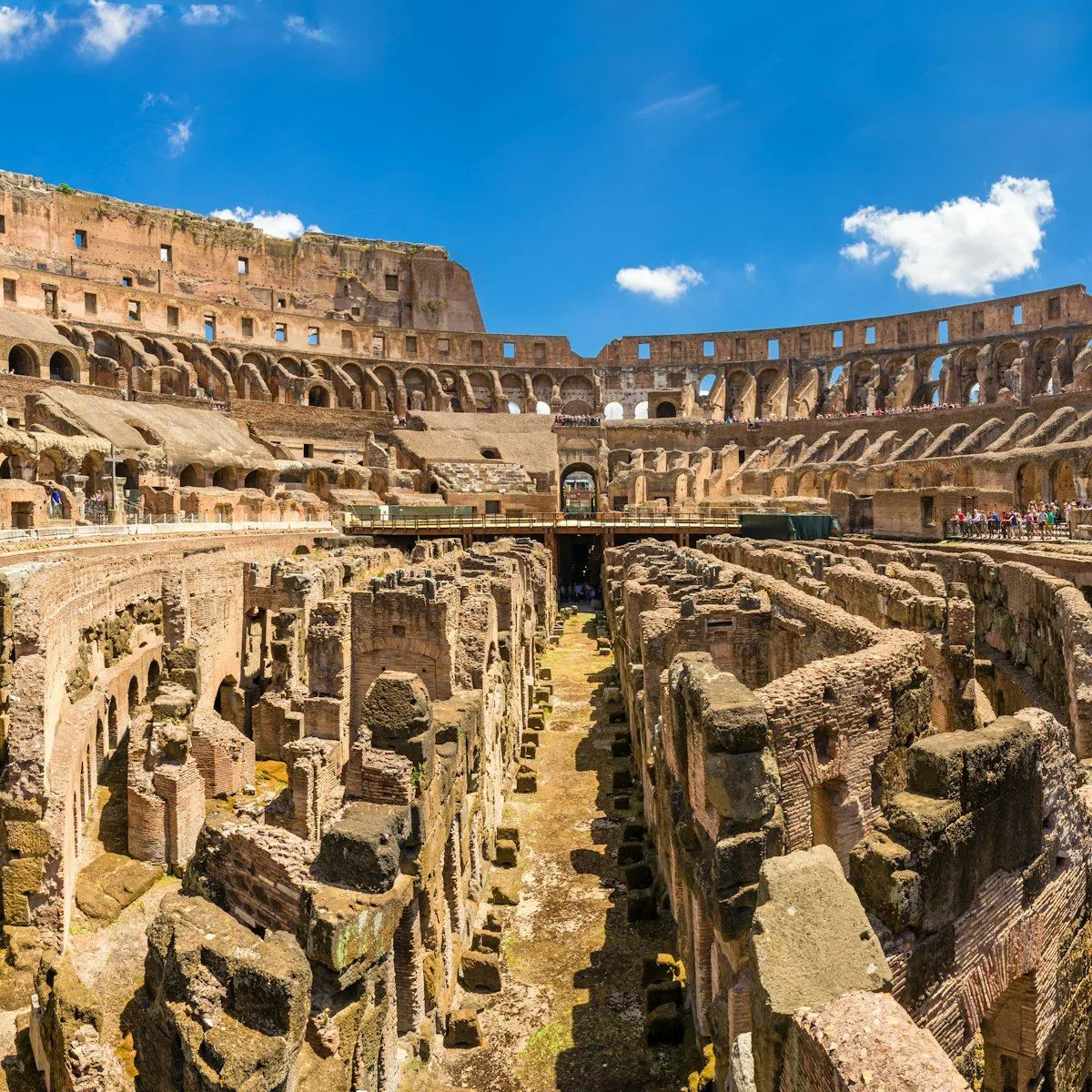 An editorial image of the interior of the Colosseum in Rome, Italy, under a clear blue sky. The image showcases the grand scale of the ancient amphitheater, including the surviving outer walls and the intricate network of the hypogeum (underground area) in the foreground.