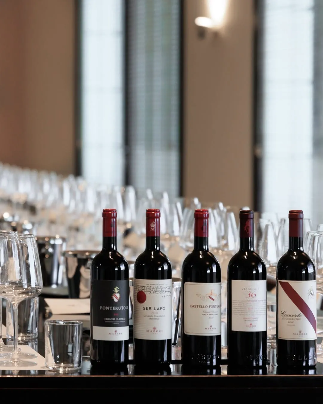 A well-composed and sharply focused image of five red wine bottles standing upright on a reflective surface, with a blurred background of numerous wine glasses. The setup suggests a wine tasting event.