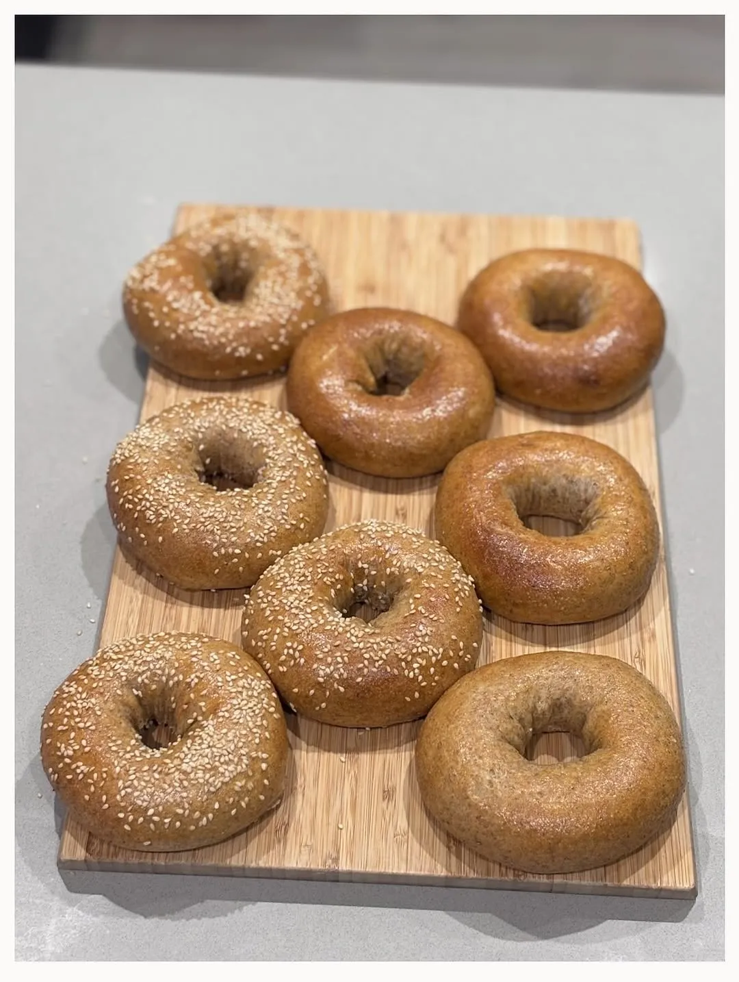 An overhead shot of several freshly baked bagels, some topped with sesame seeds, arranged on a bamboo cutting board against a neutral background.