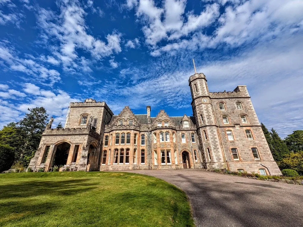An impressive, well-composed photograph of a historic stone castle under a bright blue sky with white clouds. The wide-angle lens effectively captures the scale of the building and the expansive sky.