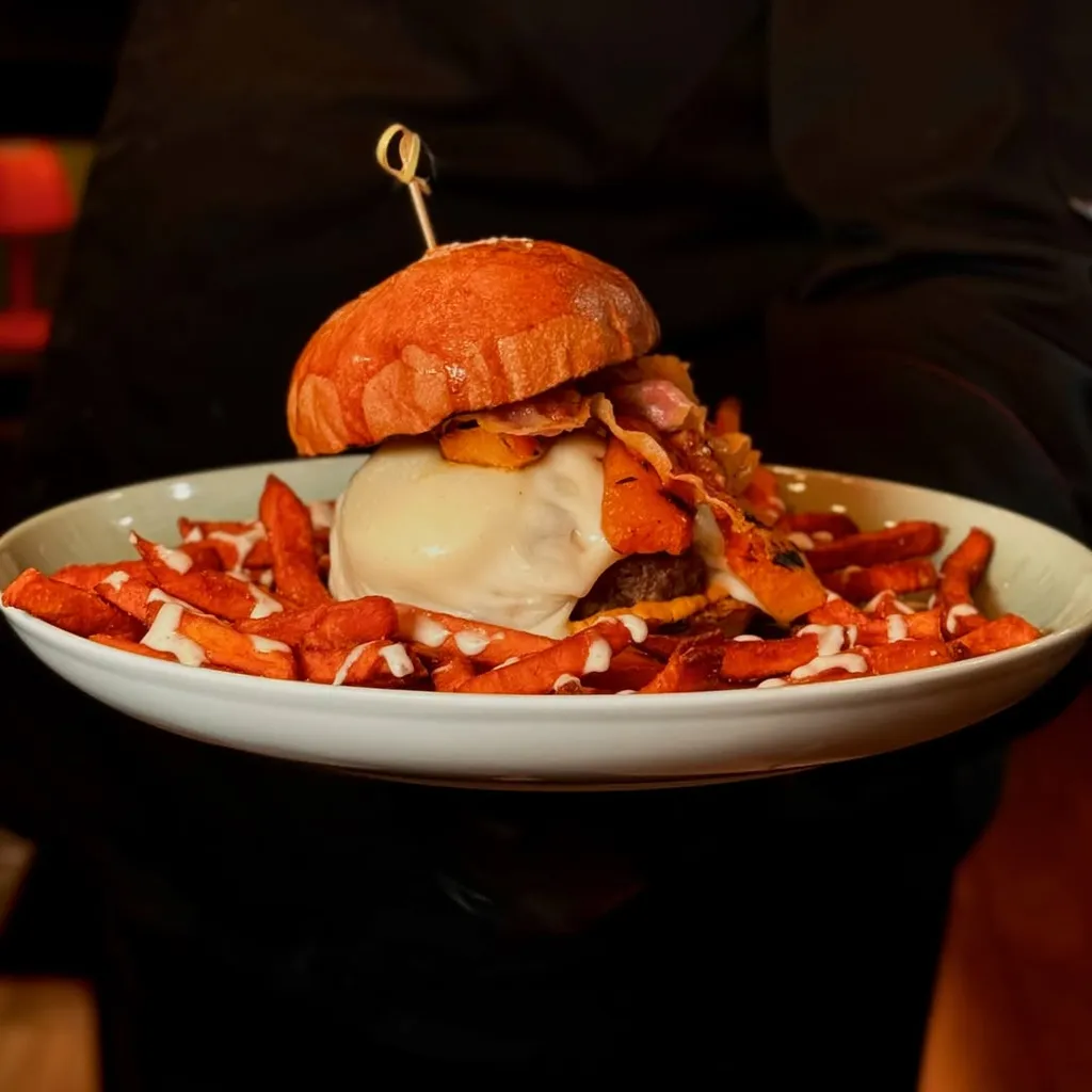 A professionally taken close-up image of a large burger with sweet potato fries, presented on a white plate and held by a person in a dark uniform. The background is dark and out of focus, highlighting the food.