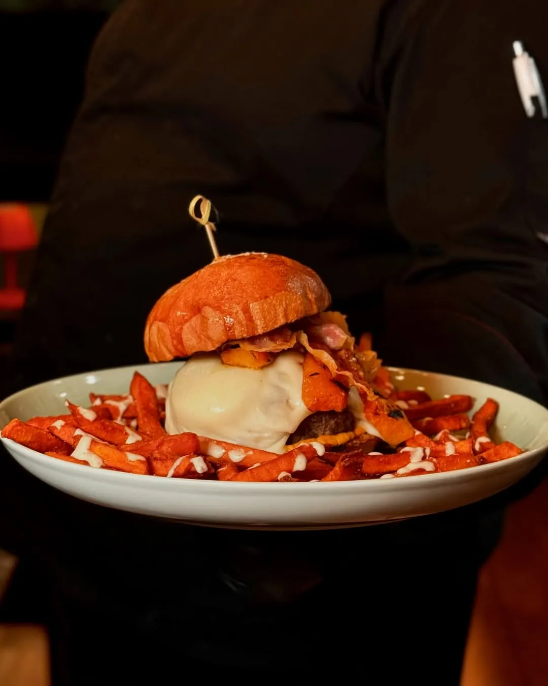 A professionally taken close-up image of a large burger with sweet potato fries, presented on a white plate and held by a person in a dark uniform. The background is dark and out of focus, highlighting the food.