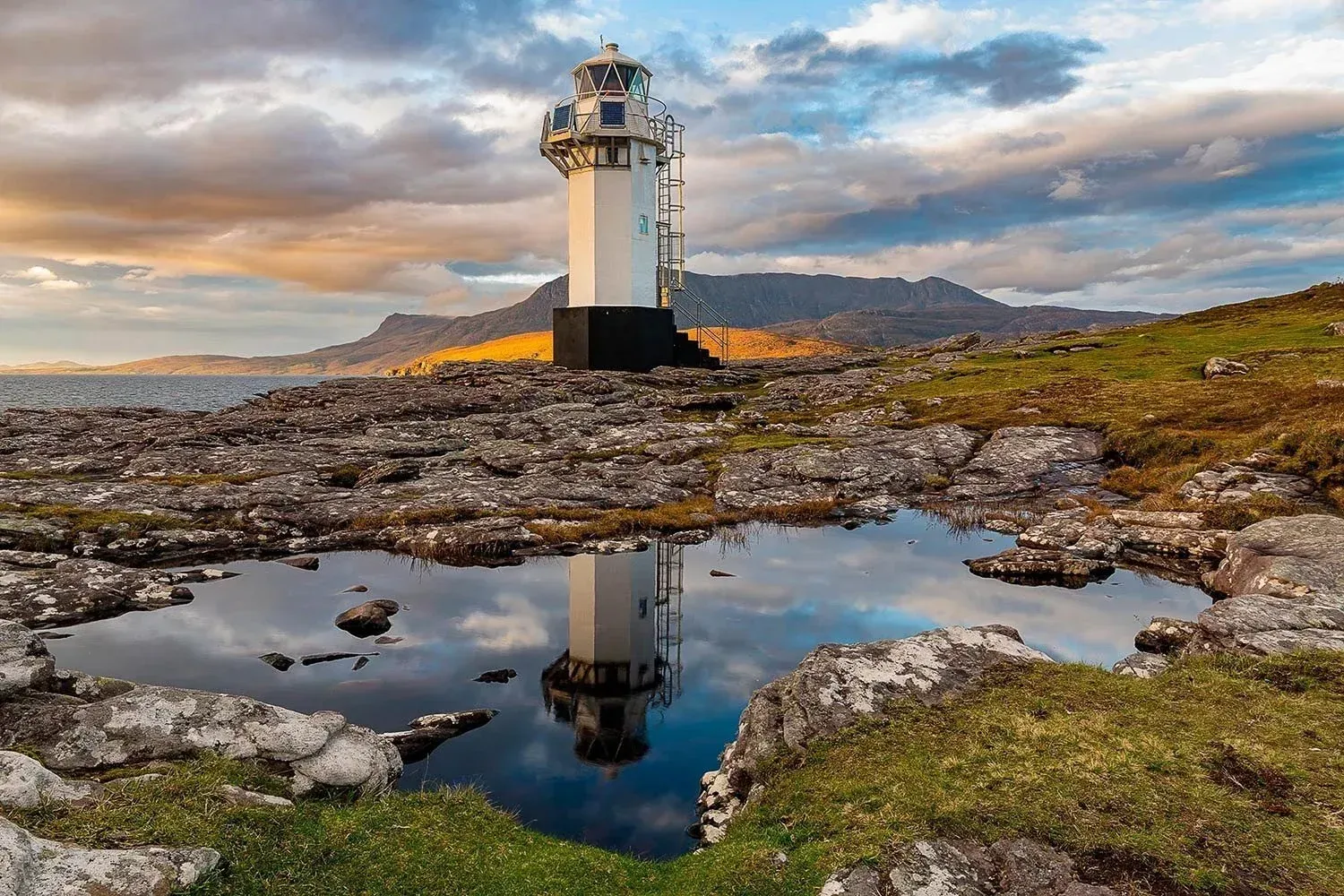 An editorial quality image of a lighthouse on a rocky coast with a reflection in a calm pool of water, set against a dramatic sky and distant mountains.