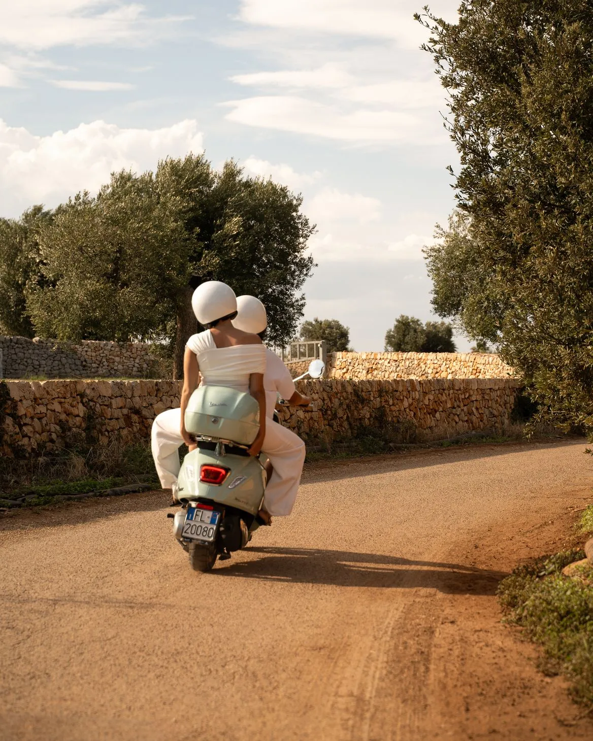 A rear view of two individuals in white clothing and helmets riding a light green Vespa scooter down a sun-drenched dirt road. The scene is set against a backdrop of traditional stone walls, olive trees, and a partly cloudy sky, evoking a sense of tranquil, rural travel.