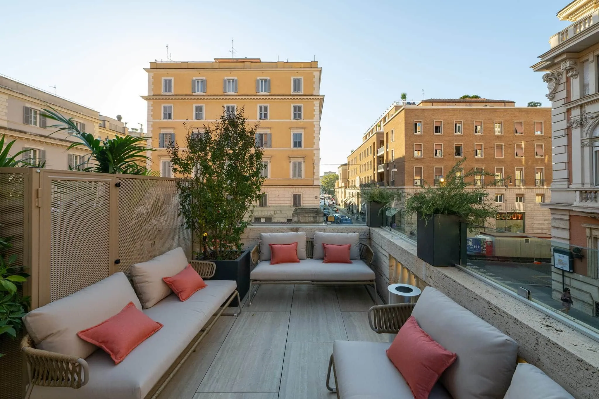 An inviting and well-composed image of a modern rooftop terrace overlooking a city street with traditional European architecture. The terrace features contemporary outdoor furniture with orange cushions and potted plants, creating an appealing urban oasis.