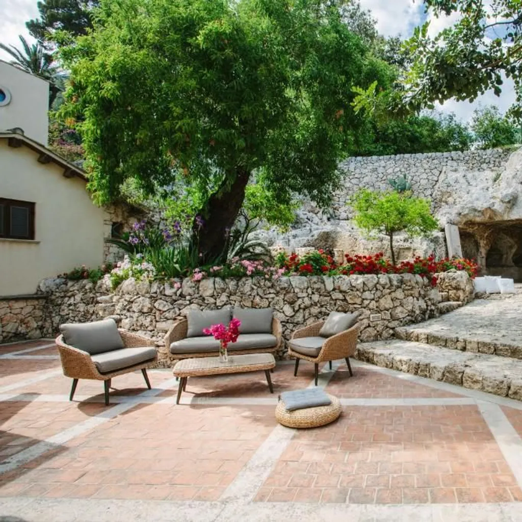 The image is well-composed, showcasing a beautiful outdoor patio area with comfortable seating, lush greenery, and unique cave-like stone architecture in the background. The natural lighting is excellent, providing an even exposure across the scene without harsh shadows or overblown highlights. The image is very sharp, with clear details visible in the furniture, stonework, and foliage. Colors appear natural and vibrant, contributing to a high degree of photo-realism. All key elements, especially the seating arrangement, are crisply in focus.