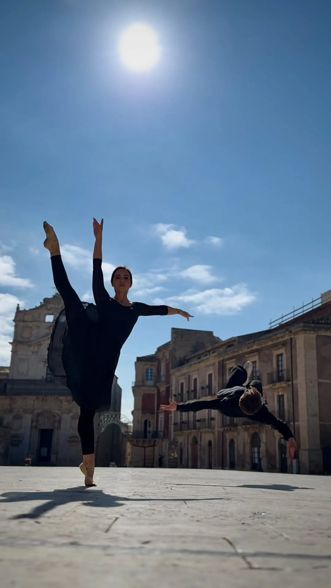 An outdoor shot capturing two dancers in black costumes performing in an urban setting under a clear blue sky. One dancer is posed with a raised leg, while the other is captured mid-air in a dynamic leap, with historic stone buildings forming the background.