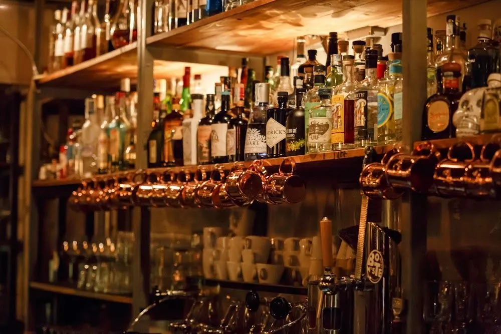A warm and inviting view inside a bar, featuring a shelf filled with various liquor bottles, a row of gleaming copper mugs hanging below, and the top of beer taps visible in the foreground. The lighting creates an atmospheric ambiance.