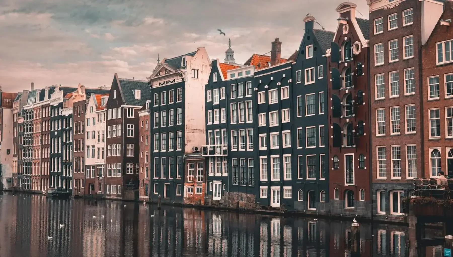 An image featuring a row of traditional canal houses, likely in a European city, with their reflections visible in the water below. The composition effectively uses the architectural elements and their mirrored images to create a visually appealing scene under an overcast sky.