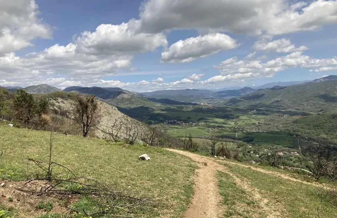 An expansive outdoor landscape scene featuring a dirt path winding through green grassy hills in the foreground, leading towards a vast valley with rolling mountains under a partly cloudy blue sky. The image captures a serene natural environment.