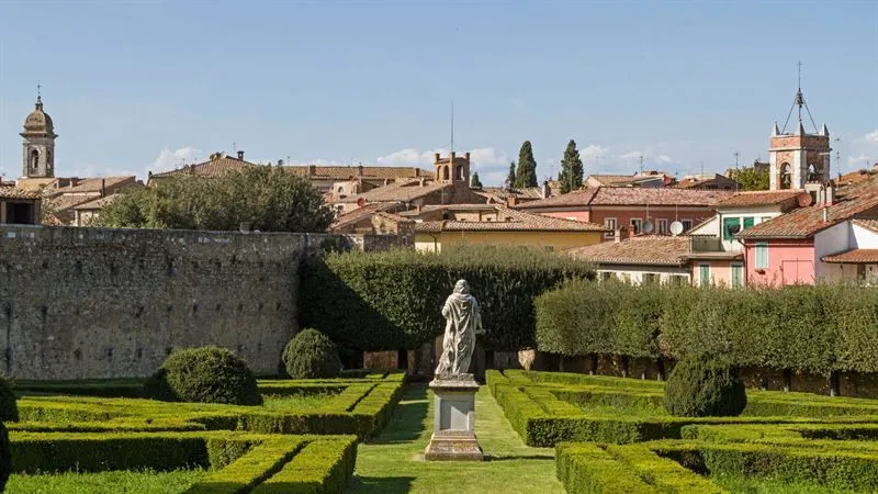 An outdoor shot of a classic Italian garden featuring manicured hedges, a central statue, and a historic town with terracotta roofs in the background under a clear sky.