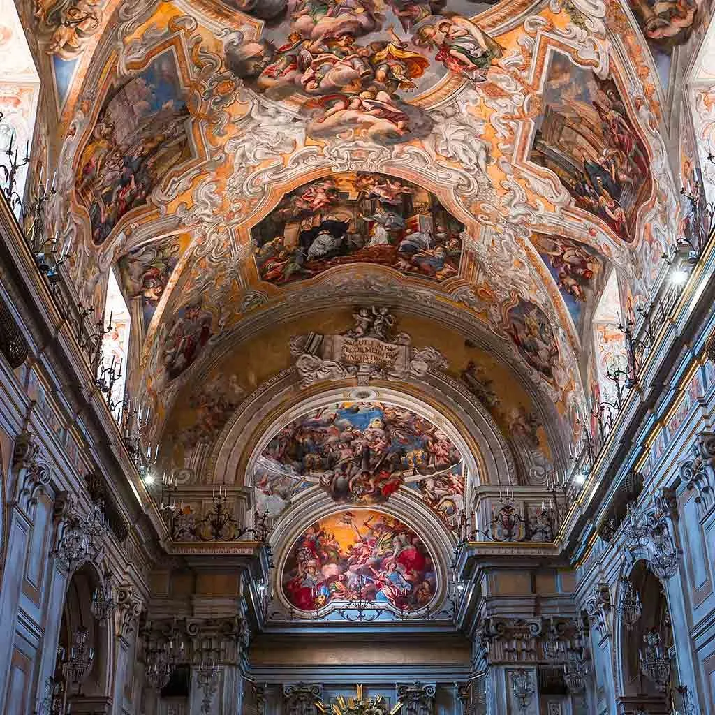 This image showcases the magnificent interior of a baroque church or cathedral, focusing on its ornate ceiling frescoes and intricate architectural details. The composition is excellent, capturing the grandeur and depth of the space effectively. The image is very sharp, ensuring that the elaborate paintings and stucco work are clearly visible, and the key elements are well in focus. The colors are rich and appear accurate, enhancing the artistic beauty of the frescoes. It is highly photo-realistic, providing a faithful representation of the interior. While generally well-lit, some peripheral areas of the lower architecture are slightly dimmer, which is common for such large indoor spaces.