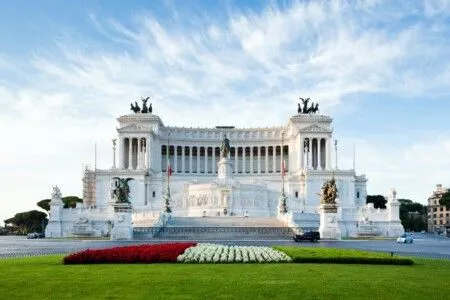 A majestic view of the Altare della Patria (Monument to Victor Emmanuel II) in Rome, Italy, showcasing its grand white marble architecture under a clear blue sky with fluffy clouds, with vibrant red and white flowerbeds and green grass in the foreground.