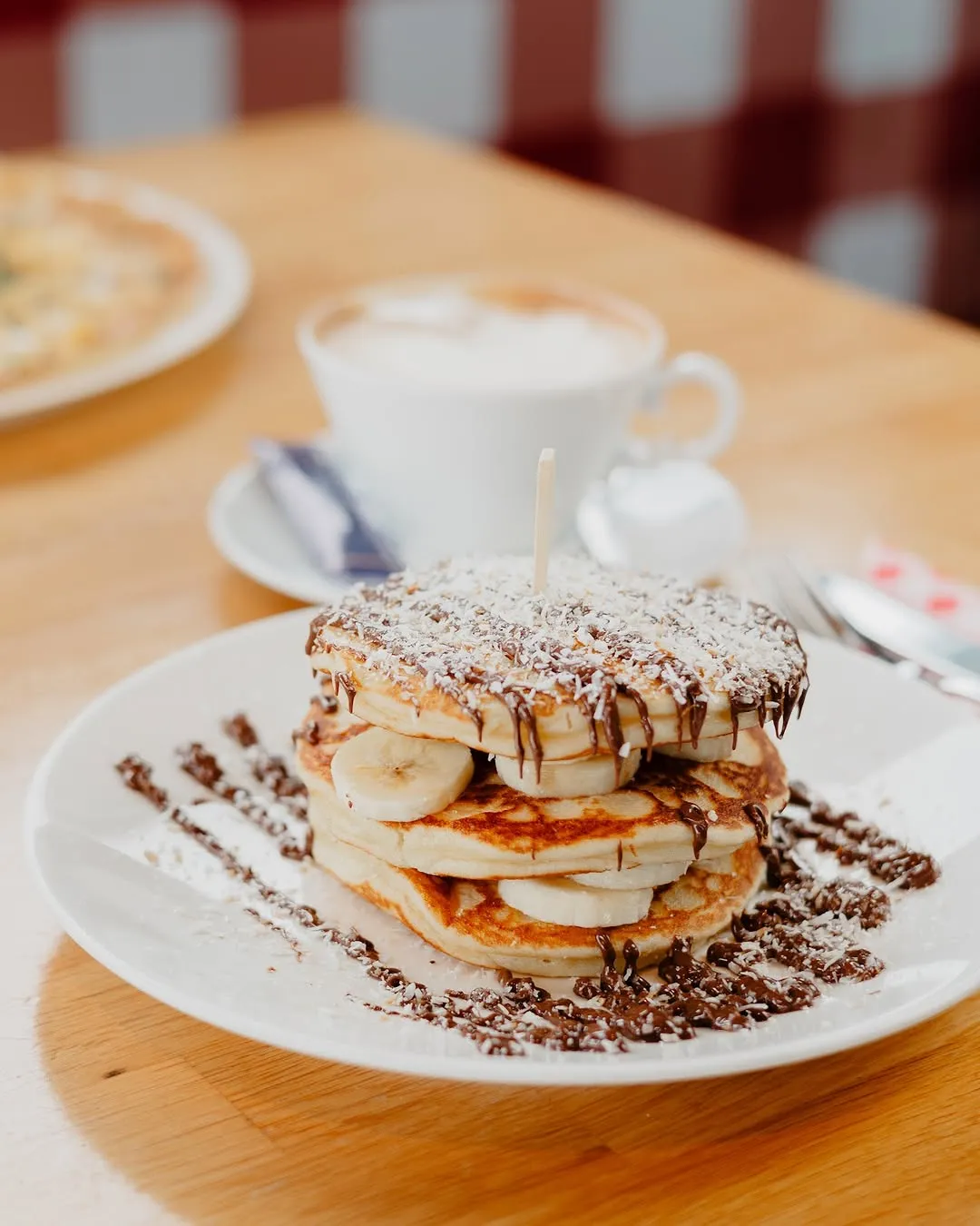 A professionally taken close-up image of a stack of pancakes with banana, chocolate drizzle, and shredded coconut, accompanied by a cup of coffee in the background. The image is well-composed and highlights the food appealingly.