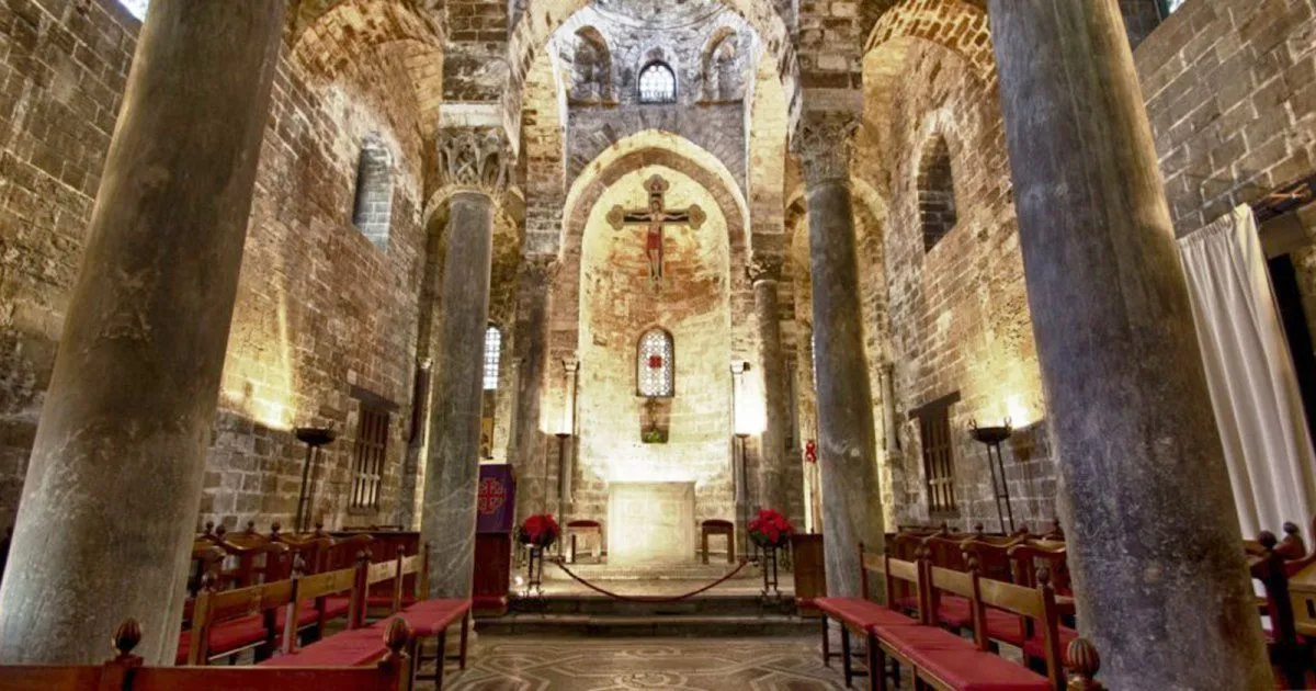 An interior shot of an old, brick-walled church or chapel, featuring a central altar with a crucifix above it, flanked by large stone columns and rows of wooden pews.