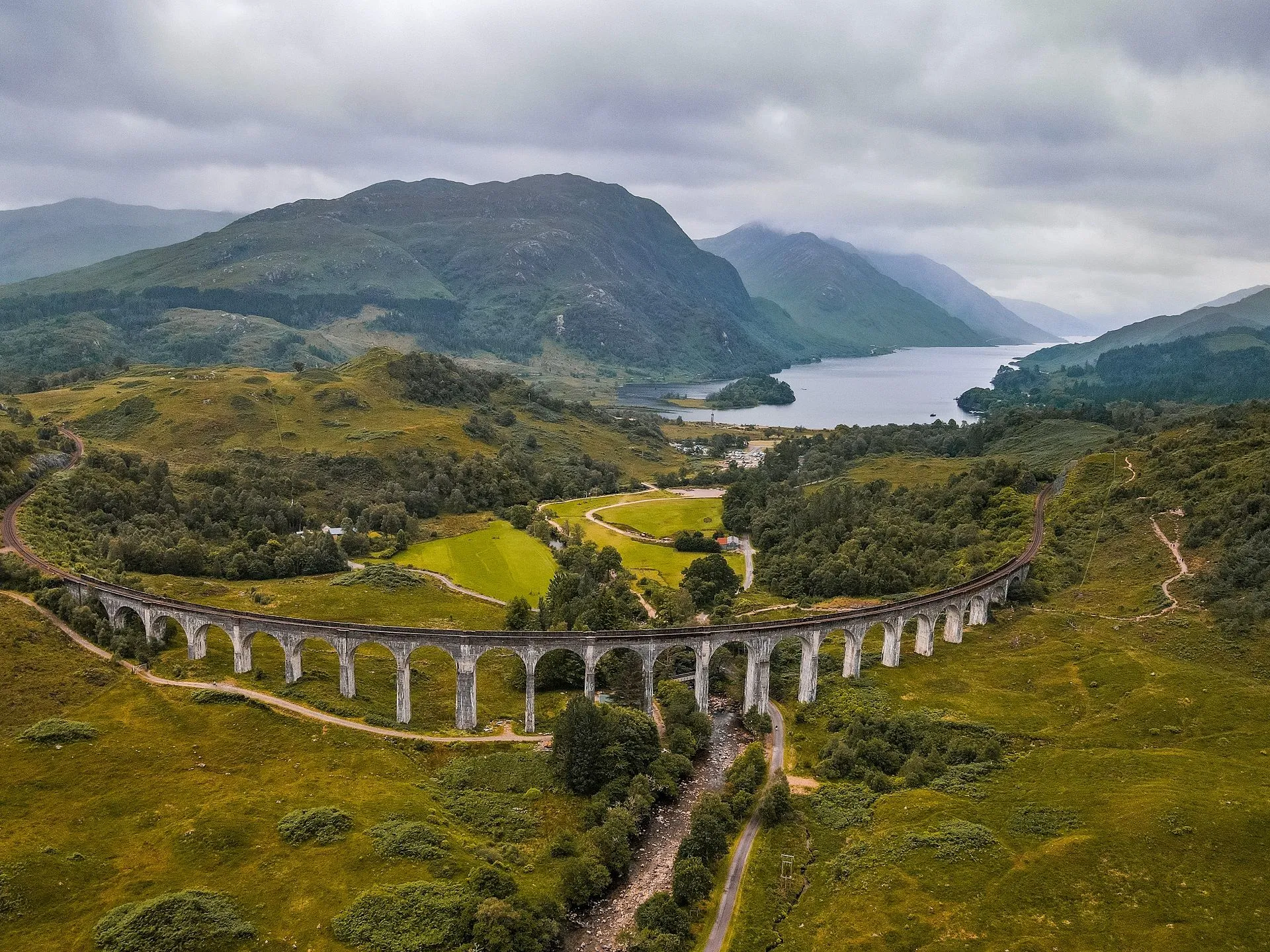 An aerial view of a majestic viaduct curving through a lush green valley, flanked by large mountains and a serene loch under an overcast sky. The composition is strong, using the viaduct as a leading line towards the background scenery.