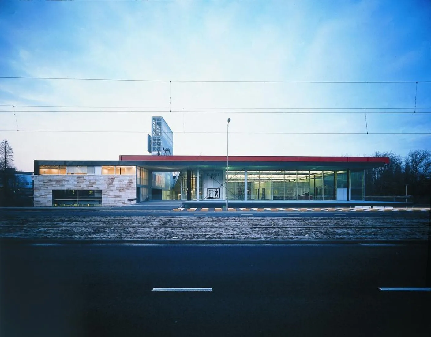 An editorial photograph featuring a modern building with a red roof, glass facades, and stone elements, viewed from across a street with tram lines. The sky is overcast, and the overall lighting is soft and flat.