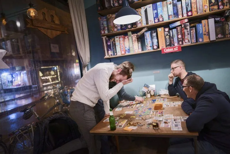 The image effectively captures a lively moment of friends playing a board game in a cafe or bar setting. The composition is strong, centering on the interaction between the players and the game itself, with board game shelves providing relevant background context. The image is highly photo-realistic and the main subjects, especially the laughing individual and the game pieces, are sharp and in focus. While the lighting is generally adequate, some areas appear a bit dim, which slightly impacts the overall brightness and clarity. The colors are natural for the indoor lighting conditions.