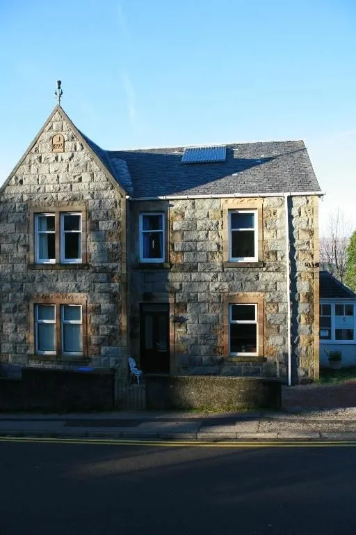 A stone house with a prominent gable and several windows, partially illuminated by direct sunlight and partially cast in deep shadow.