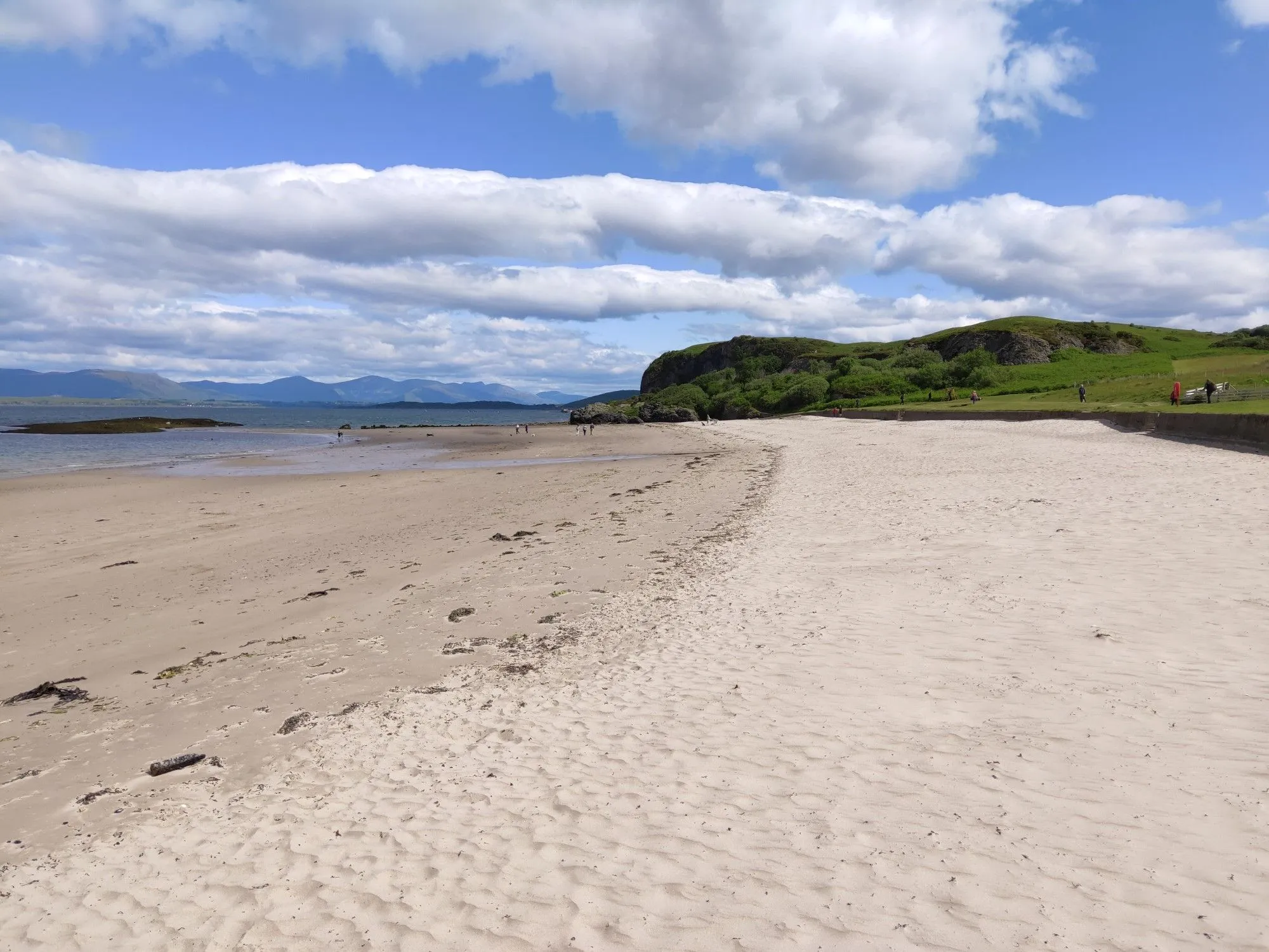 An inviting outdoor shot of a sandy beach curving along a coastline under a bright blue sky with white clouds. Distant mountains and a green, rocky headland frame the scene.