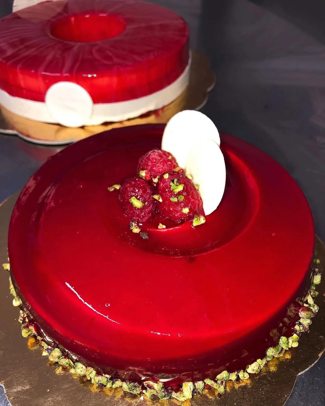 A glossy red glazed cake topped with fresh raspberries and white chocolate, with another similar cake out of focus in the background.