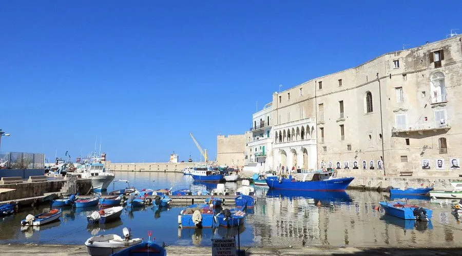An editorial image showcasing a bustling harbor with numerous boats docked in calm water, set against a backdrop of historic buildings and a clear blue sky. The scene is brightly lit by natural sunlight, highlighting the architectural details of the waterfront structures and the various vessels.