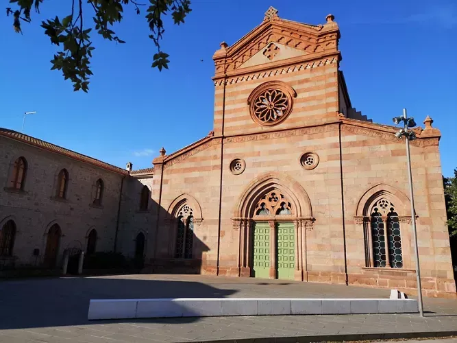 An outdoor shot of a historic church building with a distinctive reddish stone facade and green doors under a clear blue sky. Tree branches are visible in the upper left corner, and a white barrier is in the foreground.