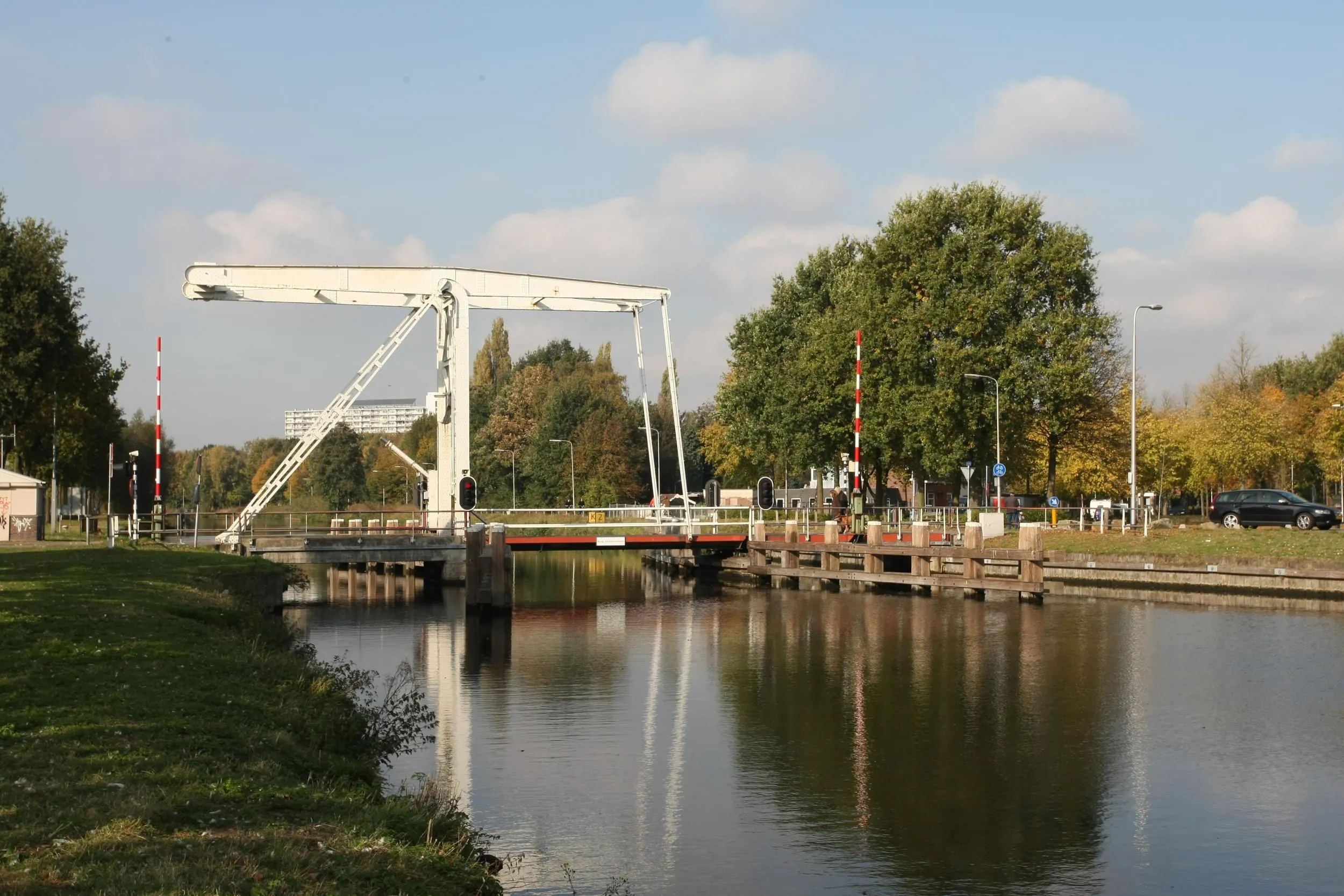 A clear and well-lit photograph of a white drawbridge spanning a canal, surrounded by lush green trees, with reflections visible on the water's surface under a bright, partly cloudy sky.