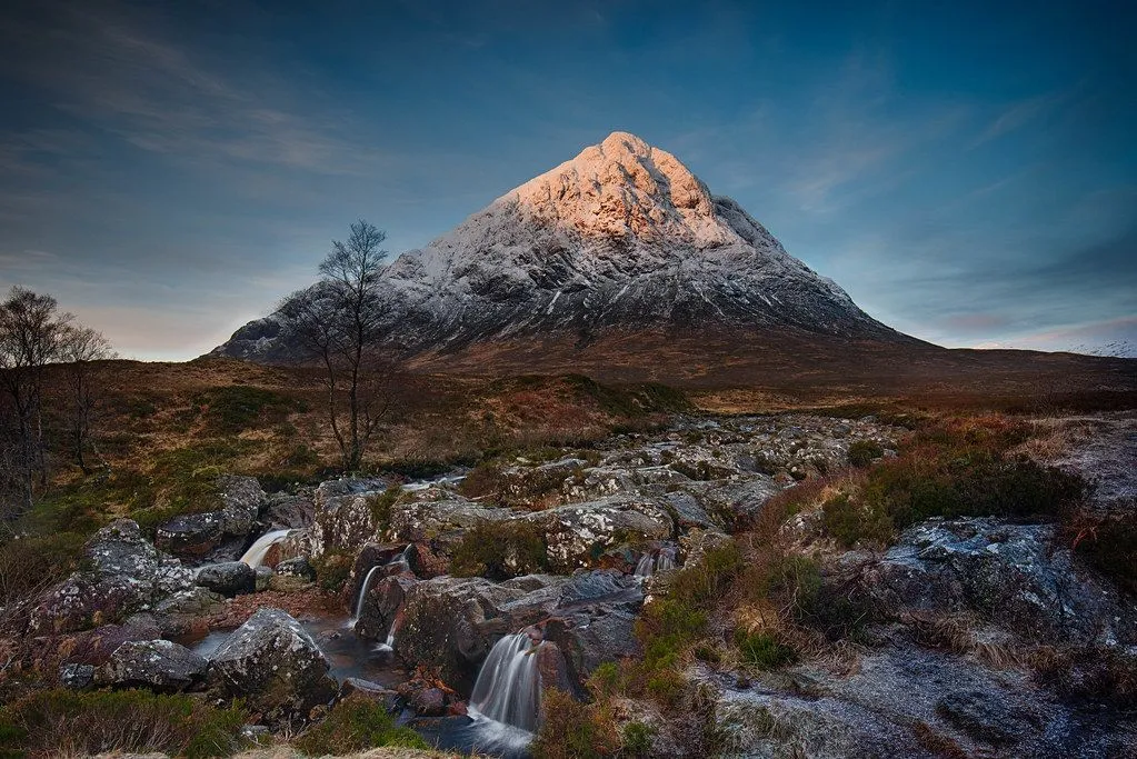 A stunning landscape photograph featuring a snow-capped mountain illuminated by warm, golden light, possibly at sunrise or sunset. A river with small waterfalls flows in the rocky foreground, adding depth and visual interest. A lone, bare tree stands to the left of the mountain. The sky is a clear blue with some light clouds.