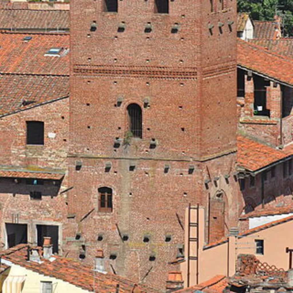 An editorial-style image of a historic brick tower, captured under clear daylight. The tower is prominently featured, showcasing its architectural details and texture against a backdrop of distant buildings and lush green trees.