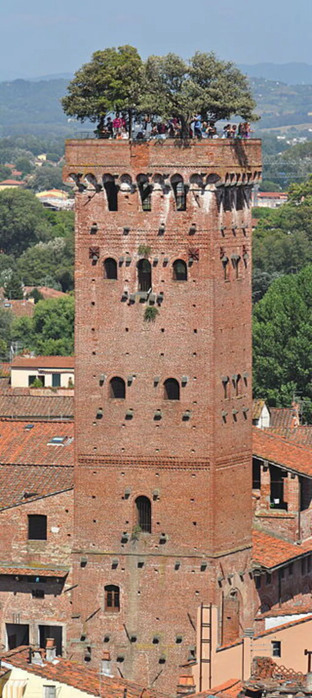 An editorial-style image of a historic brick tower, captured under clear daylight. The tower is prominently featured, showcasing its architectural details and texture against a backdrop of distant buildings and lush green trees.