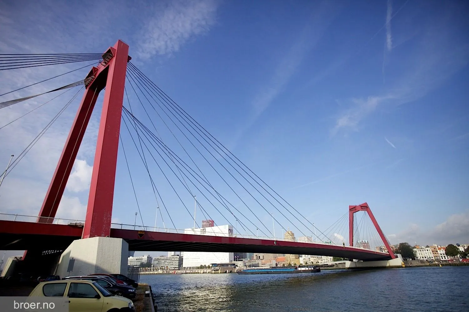 A vibrant daytime photograph featuring a distinctive red cable-stayed bridge spanning over water, set against a clear blue sky with a distant city skyline.