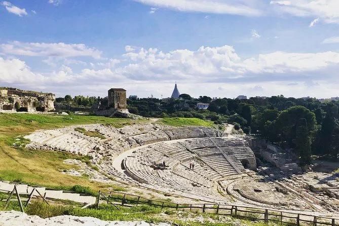 An outdoor, daytime image capturing the expansive view of an ancient Greek theater. The terraced stone seating dominates the foreground and midground, leading the eye towards the surrounding green landscape and a distant architectural structure under a bright blue sky with scattered clouds.